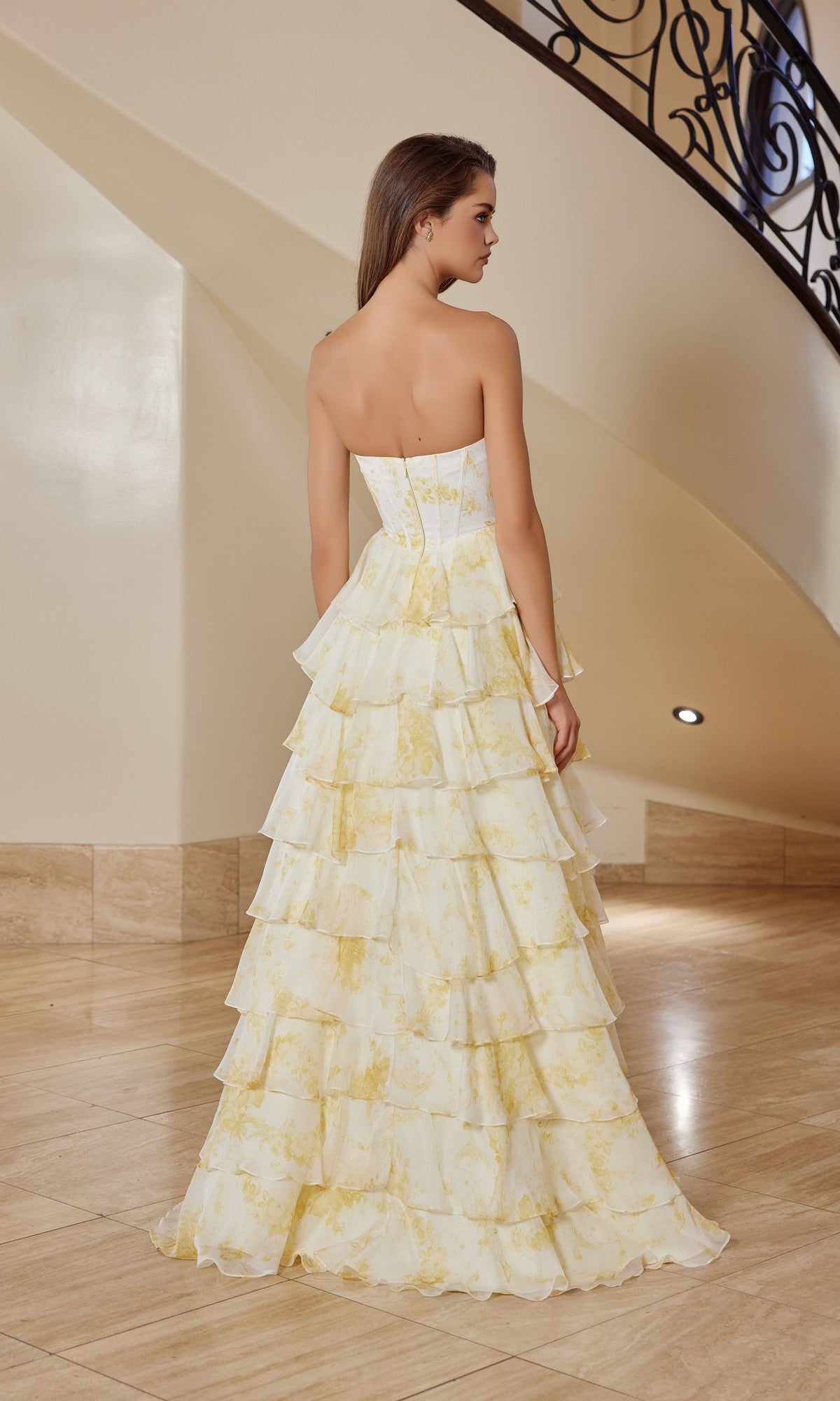 A woman stands on a marble floor near a curved staircase, wearing the Long Prom Dress: Nox Anabel T1865—a strapless floral chiffon gown with an A-line ruffle skirt—facing away from the camera.